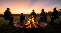 Group of friends gathered around campfire at sunset sharing stories and warmth during outdoor camping adventure in rural countryside setting.