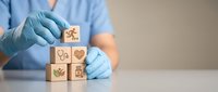 Healthcare professional in blue scrubs and gloves arranging wooden blocks with medical symbols including heart, stethoscope and health icons on white surface.