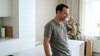 Middle-aged man with dark hair and facial stubble, wearing a gray t-shirt, drinking water from a glass in a modern kitchen with white cabinetry and natural light from windows.