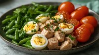 Healthy protein bowl with sliced boiled eggs, tuna chunks, fresh green beans, and cherry tomatoes on rustic ceramic plate. Perfect for clean eating meal prep.