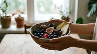 Fresh mixed berries and sliced figs in wooden bowl held by hands in bright kitchen interior with natural window light and potted plants on windowsill.