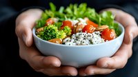 Fresh poke bowl with white rice, cherry tomatoes, broccoli and sesame seeds held in female hands against dark background. Healthy asian food concept.