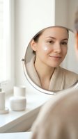 Young woman of mixed ethnicity applying skincare cream to her face while looking in a round mirror in a bright bathroom, showcasing daily beauty and wellness routine.