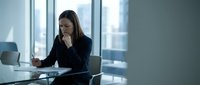 Young professional businesswoman working on laptop in modern office conference room with city view windows during daytime meeting preparation.