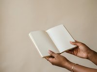 Open blank notebook held by person with dark skin tone against neutral beige background, suitable for publishing, education, writing, and creative projects.