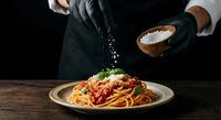 Chef hands sprinkling parmesan cheese over spaghetti with tomato sauce on wooden table in restaurant kitchen for culinary presentation.