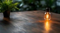 Glowing vintage filament bulb hanging over wooden table surface with soft warm light reflection and decorative plant in background, moody atmosphere.