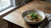 Steaming hot Asian noodle soup with fresh herbs and vegetables served in ceramic bowl on rustic wooden table by window for cozy dining atmosphere.