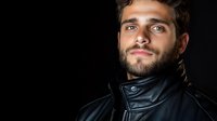 Young bearded Caucasian man in black leather jacket looking up with confident smile against dark background, dramatic studio lighting highlights facial features.