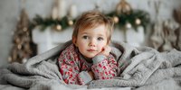 Adorable toddler wrapped in cozy gray blanket with festive Christmas decorations in background, wearing floral patterned outfit for holiday portrait.
