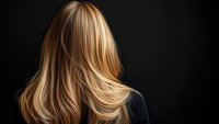 Back view of woman with flowing blonde highlighted hair against dark background, showcasing professional salon styling for hair care advertising.
