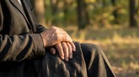 Senior man sitting peacefully in autumn park with hands resting on walking cane, enjoying golden hour sunlight filtering through trees in background.