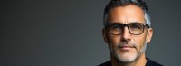 Confident mature man with gray hair and designer glasses looking directly at camera against dark background, professional headshot style portrait.