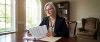 Mature woman with gray hair and glasses, Caucasian ethnicity, wearing black blazer, reviewing documents at wooden desk in home office with natural light from window, suitable for business, professional services, and corporate concepts.