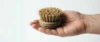 Hand holding a small round natural fiber dish brush, eco-friendly cleaning tool made from sustainable materials, isolated on white background.