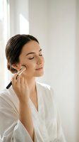 Young woman applying makeup with brush in bright bathroom wearing white robe. Natural morning skincare and beauty routine for daily self-care ritual.