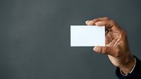 Mature male hand with medium brown skin tone holding blank white business card against neutral gray background, suitable for corporate branding and professional identity design.