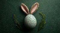 Speckled egg with handcrafted bunny ears made from pink fabric and twine, placed between fresh rosemary sprigs on dark green textured background. Creative Easter concept.