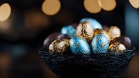 Luxury chocolate Easter eggs wrapped in blue and gold foil arranged in dark decorative nest bowl against blurred warm bokeh lights background.