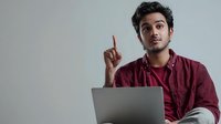 Young South Asian man in burgundy shirt raising index finger in eureka moment while sitting with laptop against neutral background. Perfect for business presentations.