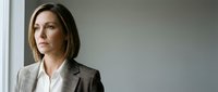 Professional businesswoman in gray suit looking thoughtfully out office window, corporate executive contemplating future business decisions and strategy.