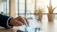 Professional businessman hand touching digital communication icons including phone, email and live chat on modern office desk with warm sunlight and plant background.