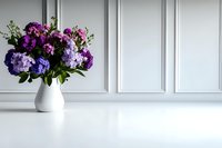 Purple and pink hydrangea flowers arranged in white ceramic vase against classic white wall paneling, creating minimalist elegant home decor composition on reflective surface.