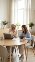 Senior businesswoman mentoring young female colleague at modern office table with laptop and tablet for professional development and workplace collaboration.