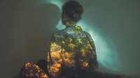Double exposure silhouette of woman merged with autumn forest trees creating artistic portrait with warm orange yellow foliage against teal background.