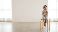 Young child, appearing to be of mixed ethnicity, sitting on a wooden stool in a bright, minimalist interior space with white walls and natural light from windows.