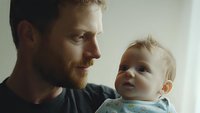 Caucasian father with ginger beard holding baby in tender moment near window. Natural light creates intimate atmosphere in emotional parenting scene.