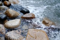 Waves crashing against smooth river rocks create dynamic water movement and natural texture, capturing the raw energy of flowing water meeting stone shoreline.