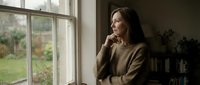 Middle-aged woman with long dark hair and olive complexion gazes thoughtfully out window in contemplative moment, suitable for editorial, lifestyle, and wellness content.