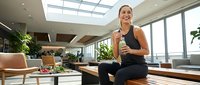 Young woman with dark hair and olive complexion, wearing black athletic wear, sitting on wooden bench in modern office gym or wellness space with plants, large windows, and contemporary furniture, holding water bottle during fitness break.