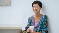 Asian woman with short dark hair enjoying morning coffee at home, wearing casual colorful layered clothing, sitting relaxed with warm smile against white wall background.