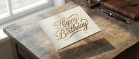 Elegant Happy Birthday greeting card with gold lettering on cream paper, placed on rustic wooden table near window with natural lighting and leather accessories.