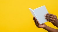 Person with dark skin tone holding open blank white book against bright yellow background, reading or studying.