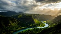 Aerial view of a winding turquoise river flowing through lush green mountains and valleys under dramatic cloudy sky with golden sunlight breaking through clouds.