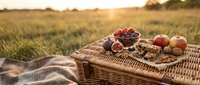 Wicker picnic basket with fresh fruits, nuts and berries on blanket in golden sunset field for outdoor dining and leisure activities.
