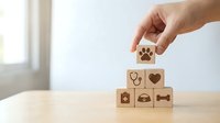 Hand placing wooden cube with paw print icon on pyramid of pet care symbols including stethoscope, heart, medical cross and bone on table.