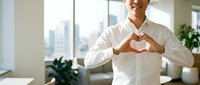 Smiling Asian businessman in white shirt making heart shape with hands in modern office, expressing care, positivity, and corporate wellness.