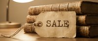 Vintage sale tag with rope attached leaning against stack of old antique books on wooden table with warm lamp light in background.