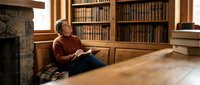 Young Caucasian man reading book in cozy library with wooden shelves and fireplace, relaxed learning atmosphere for educational content.
