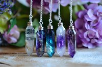 Crystal point pendants with silver chains displayed against purple hydrangea background, featuring quartz, amethyst and labradorite minerals in hexagonal cuts.
