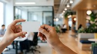 Hands holding blank white business card in modern office lobby with blurred colleagues and plants in background, suitable for corporate branding and networking concepts.