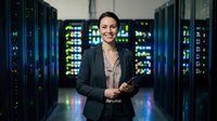 Young female IT professional with dark hair and olive complexion wearing business blazer standing confidently in data center surrounded by illuminated server racks, suitable for technology, cloud computing, and corporate business concepts.