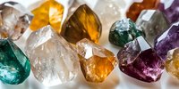 Raw healing crystals and mineral stones in various colors including amethyst, citrine, clear quartz, and emerald displaying natural formations and facets in close-up view.