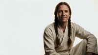 Native American man with long braids in traditional beige clothing sitting peacefully against neutral background for cultural and historical projects.