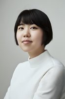 Young Asian woman with short black bob haircut wearing white turtleneck sweater looking up thoughtfully against neutral gray background in minimalist portrait style.