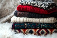 Folded knit sweaters and cozy winter clothing stacked in warm colors including red, cream, and patterned designs on soft fur background for seasonal fashion and wardrobe styling.